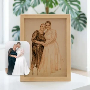 Un portret pirogravat 30x40 cu două femei îmbrățișându-se este expus pe o masă, cu fotografia originală în față și o plantă cu frunze verzi în fundal, evidențiind această pictură specială după poză.